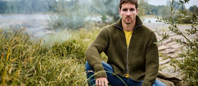 Ein Mann sitzt im Grünen und trägt eine olivgrüne Strickjacke mit Reißverschluss über einem gelben T-Shirt und einer blauen Jeans.