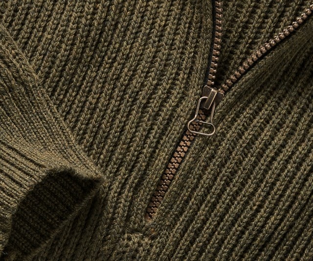 Das Bild zeigt eine Nahaufnahme einer grob gestrickten, olivgrünen Strickjacke mit einem metallischen Reißverschluss.