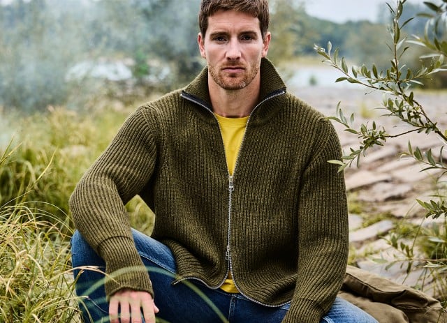 Ein Mann sitzt in der Natur am Ufer, trägt eine olivgrüne Strickjacke über einem gelben T-Shirt und blickt ruhig in die Kamera.