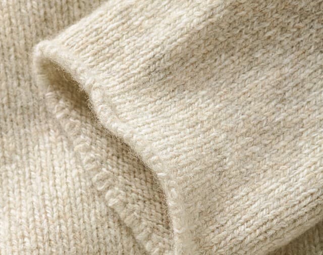 Das Bild zeigt eine Nahaufnahme eines weichen, grob gestrickten Wollstoffs in hellem Beigeton, der eine warme und natürliche Textur erkennen lässt.