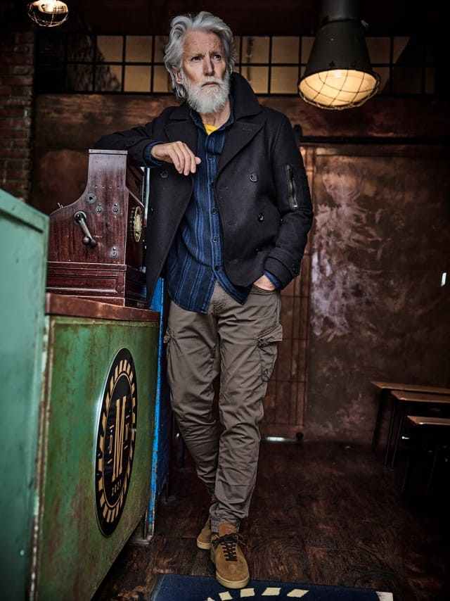 Ein Mann trägt eine dunkelblaue Jacke über einem blauen Hemd mit gelbem T-Shirt, dazu beige Cargohosen und braune Wildlederschuhe – ein lässiger, stilvoller Freizeitlook.