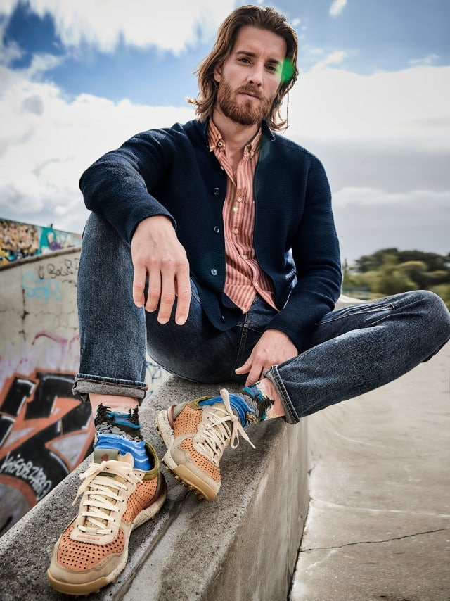 Ein Mann trägt ein lässiges Outfit aus dunkelblauer Strickjacke, rot-weiß gestreiftem Hemd, aufgekrempelter Jeans und beigen Sneakern mit bunten Socken – ein moderner, entspannter Freizeitlook.