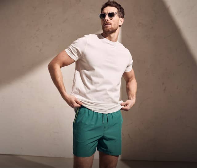 Auf dem Bild steht eine Person mit Sonnenbrille, weißem T‑Shirt und grünen Shorts vor einer hellen Wand, während schräg einfallendes Licht Schatten auf den Hintergrund wirft.