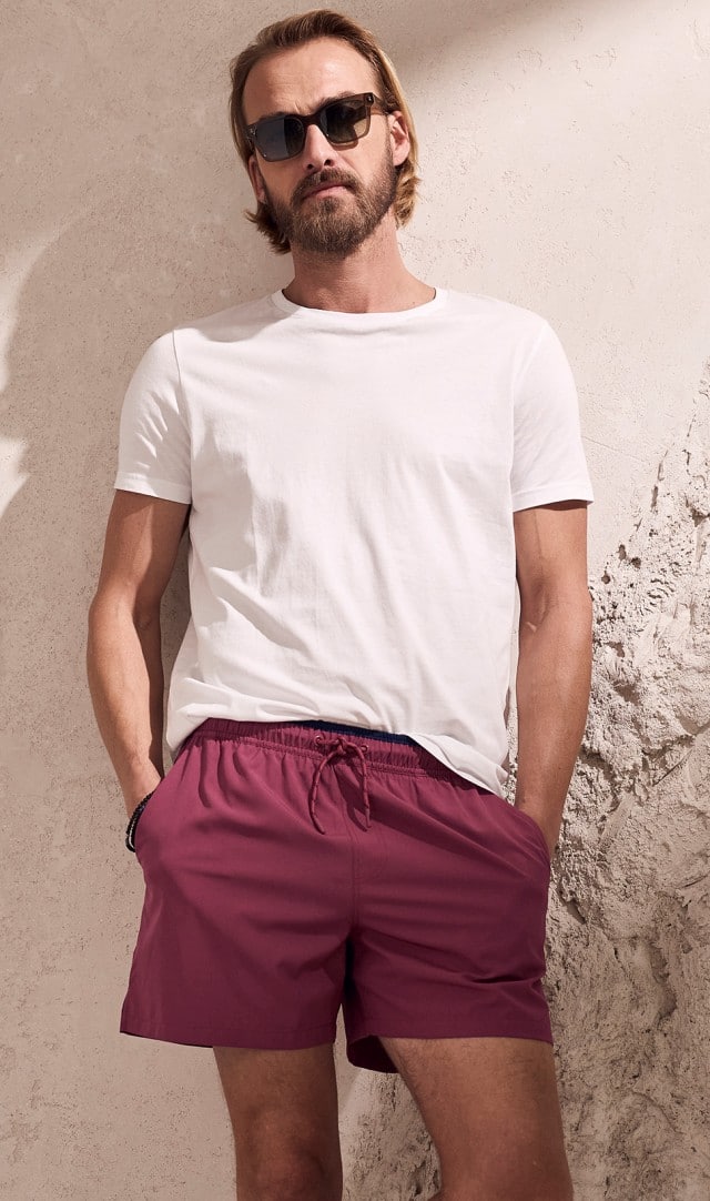 Auf dem Bild lehnt eine Person mit Sonnenbrille, weißem T‑Shirt und roten Shorts entspannt an einer hellen, strukturierten Wand.