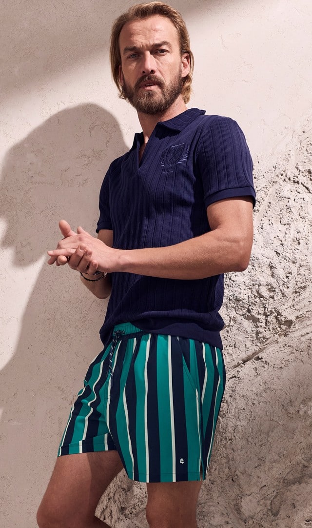 Auf dem Bild steht ein Mann in einem dunkelblauen Poloshirt und grün‑gestreiften Shorts mit verschränkten Händen vor einer hellen, strukturierten Wand.