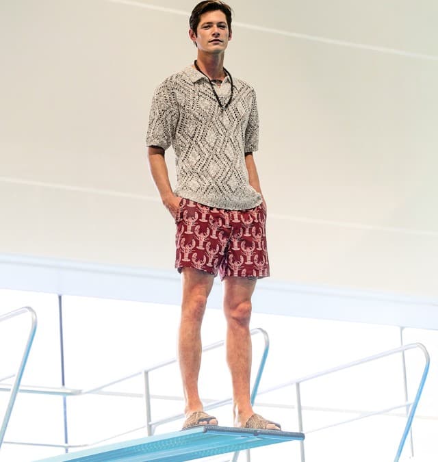 Auf dem Bild steht eine Person auf einem Sprungbrett, trägt ein gemustertes Kurzarmhemd und rote Shorts und blickt in die Ferne.