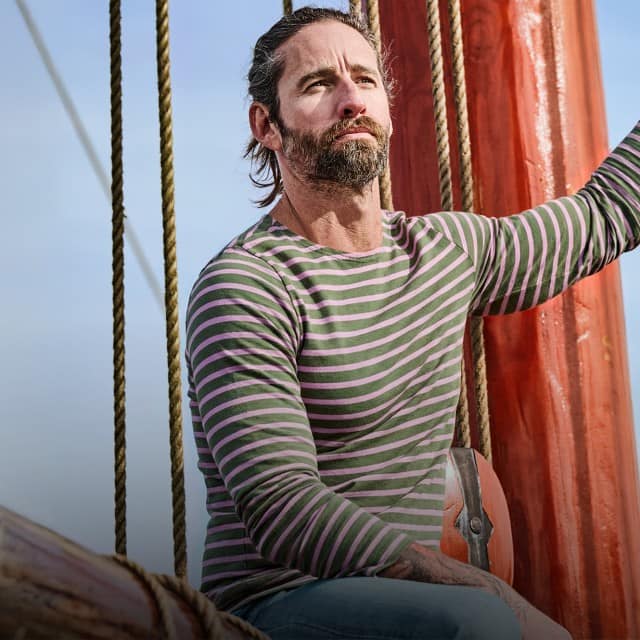 Ein Mann sitzt auf einem Segelschiff und trägt ein langärmliges, grün‑rosa gestreiftes Shirt, während er sich an einem der Taue festhält.