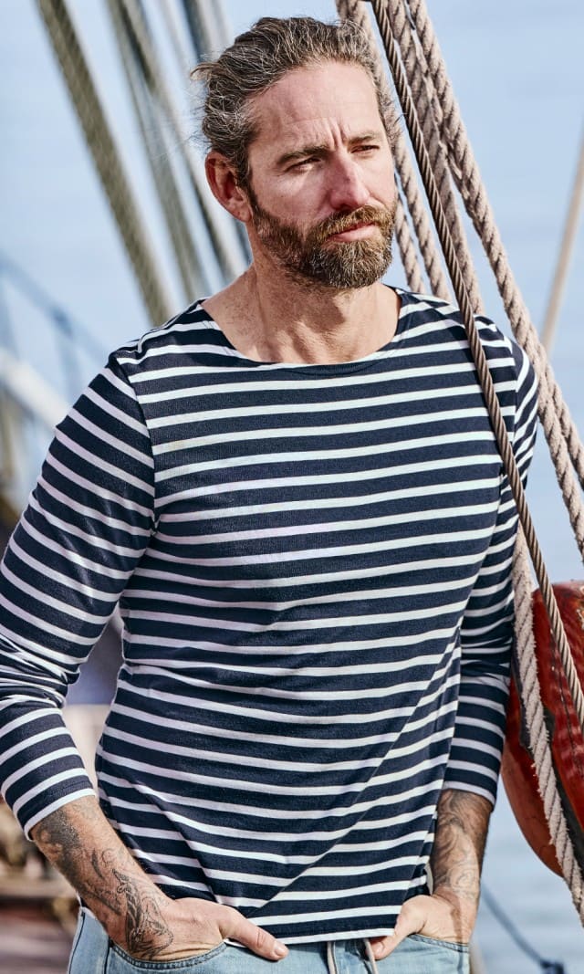 Ein Mann trägt ein langärmliges Shirt mit navy‑weißen Streifen und eine hellblaue Jeans, während er auf einem Segelschiff steht.
