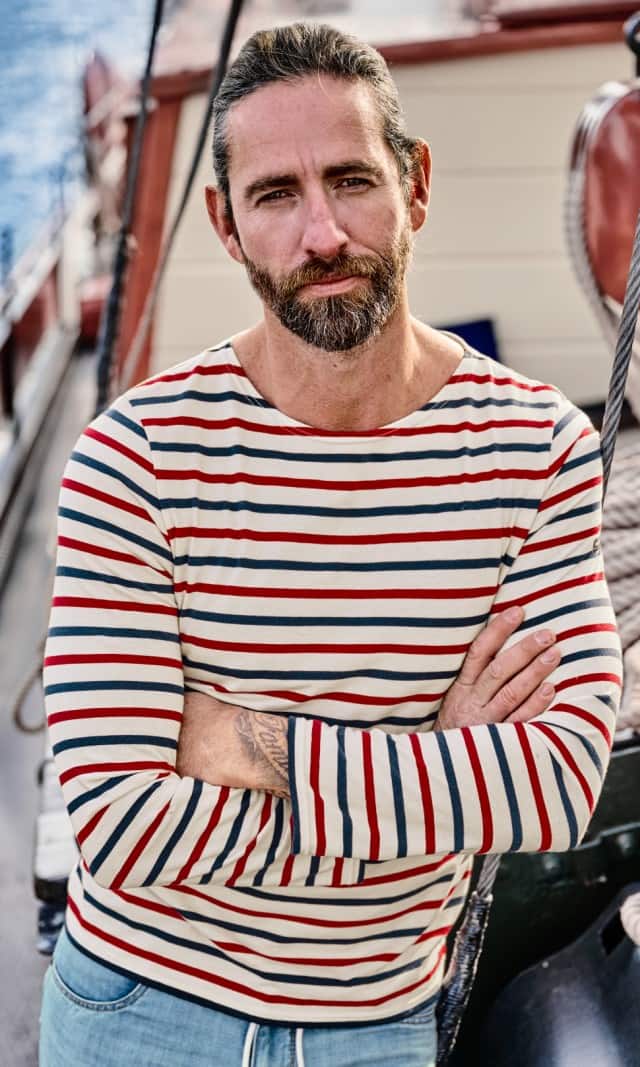 Ein Mann trägt ein rot-weiß blau gestreiftes langärmeliges Shirt  und eine hellblaue Jeans, während er auf einem Segelschiff steht.