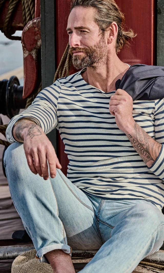 Ein Mann trägt ein langärmliges Shirt blau weißen Streifen und eine hellblaue Jeans, während er auf einem Segelschiff sitzt.