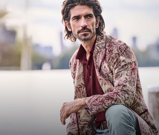 Ein Mann sitzt am Wasser und trägt eine gemusterte beige‑rote Jacke über einem bordeauxfarbenen Shirt sowie eine hellgraue Jeans.