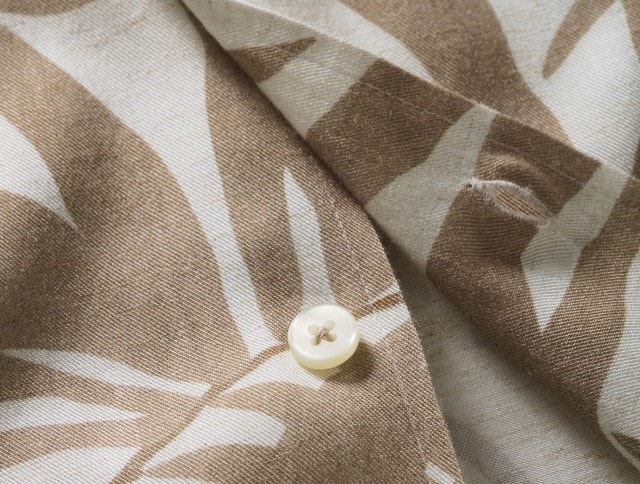Eine Nahaufnahme zeigt einen beige-weiß gemusterten Stoff mit einem Knopf, der die feine Textur und das sommerliche Design betont.
