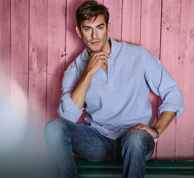 Ein Mann bekleidet mit einem blauen Leinenhemd sitzt entspannt vor einer Holzwand