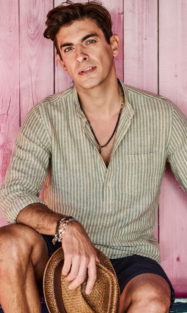 Ein Mann trägt ein grün gestreiftes Leinenhemd, dazu eine dunkelblaue Shorts und sitzt auf einer Bank,