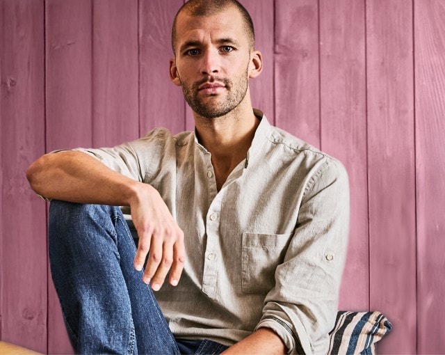 Das Bild zeigt einen Mann mit kurzgeschnittenen Haaren und Bart, der in lässiger Kleidung auf einem Sofa vor einer pink gestrichenen Holzwand sitzt.