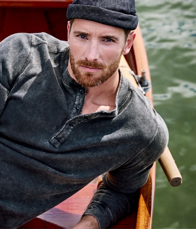 Ein Mann sitzt in einem Ruderboot und trägt ein dunkelgraues, langärmliges Henley-Shirt, eine graue Hose und eine dunkle Schirmmütze.