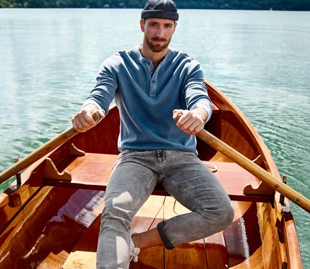 Auf dem Bild sieht man links einen Mann, der in einem Ruderboot sitzt und ein blaues Henley-Langarmshirt trägt, während rechts das gleiche Shirt einzeln abgebildet ist.