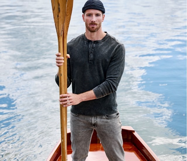 Auf dem Bild ist links ein Mann zu sehen, der in einem Ruderboot steht und ein dunkles, langärmliges Henley-Shirt sowie eine Mütze trägt; rechts davon ist das gleiche schwarze Henley-Shirt einzeln abgebildet.
