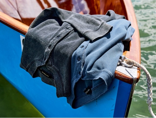 Auf dem Bild liegen zwei Henley-Langarmshirts, eines in schwarz und eines in blau, nebeneinander über den Rand eines blauen Ruderbootes am Wasser.