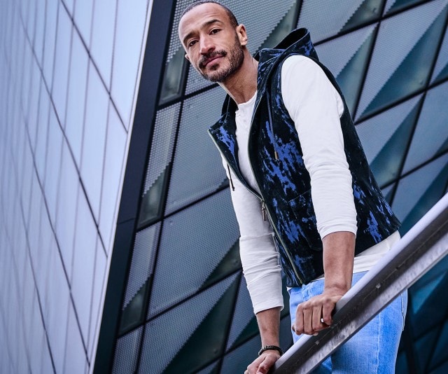Ein Mann steht an einem Geländer vor einem modernen Glasgebäude, trägt eine weiße Langarmshirt und eine dunkle, gemusterte Weste und blickt in die Kamera.
