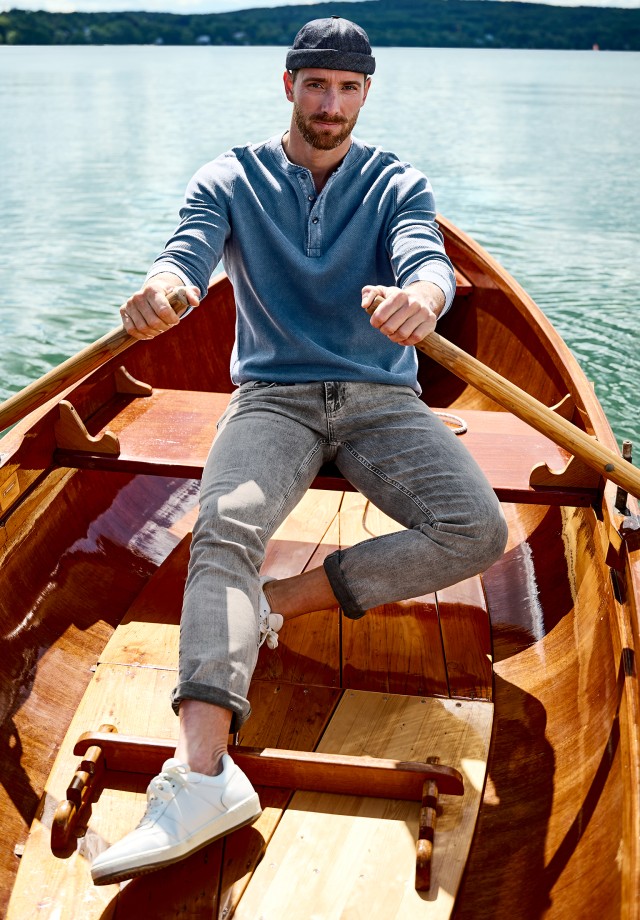 Auf dem Bild sieht man links einen Mann, der in einem Ruderboot sitzt und ein blaues Henley-Langarmshirt trägt, während rechts das gleiche Shirt einzeln abgebildet ist.