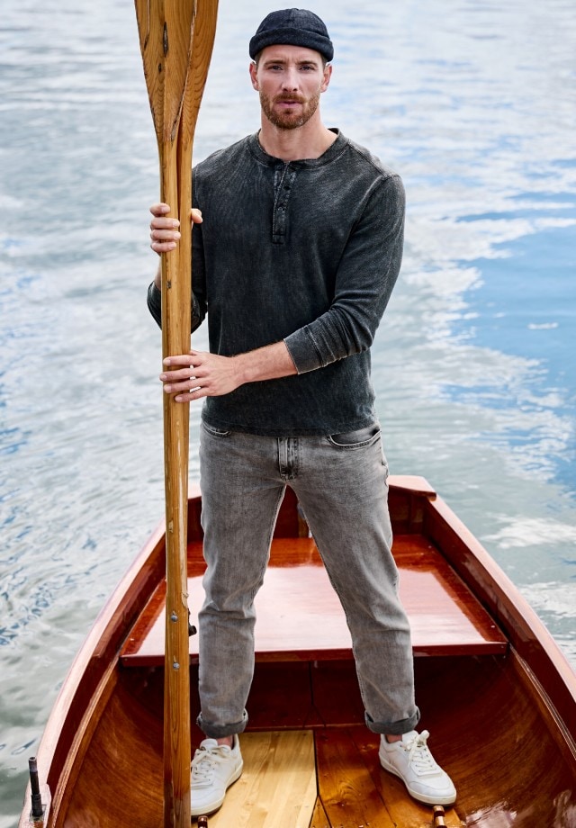 Auf dem Bild sieht man links einen Mann, der in einem Ruderboot sitzt und ein schwarzes Henley-Langarmshirt trägt, während rechts das gleiche Shirt einzeln abgebildet ist.