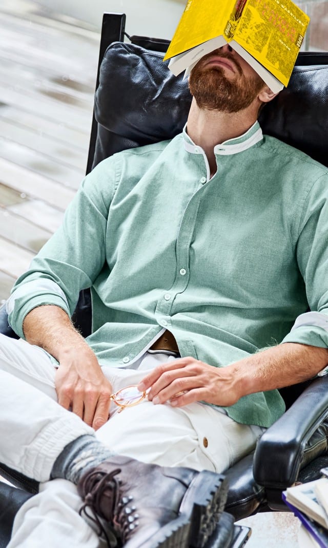 Der Mann trägt ein mintgrünes Stehkragenhemd, weiße Hose und dunkle Boots, während er entspannt in einem Sessel liegt.