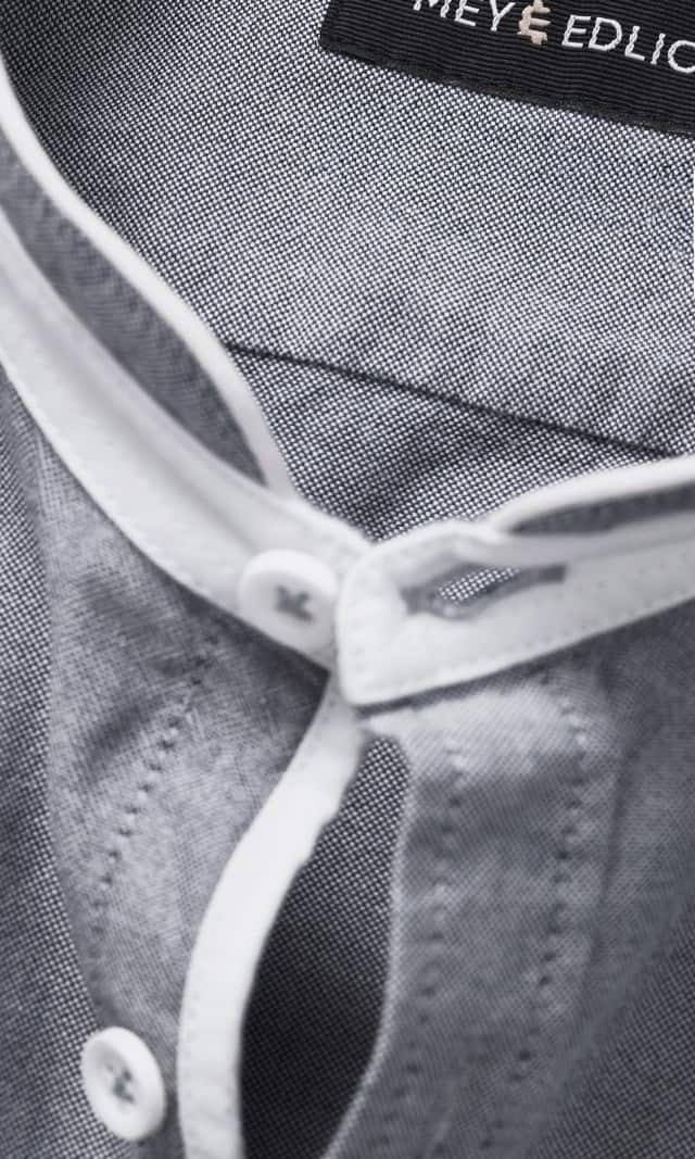 Der Mann trägt ein graues Stehkragenhemd mit hochgekrempelten Ärmeln sowie eine weiße Hose.