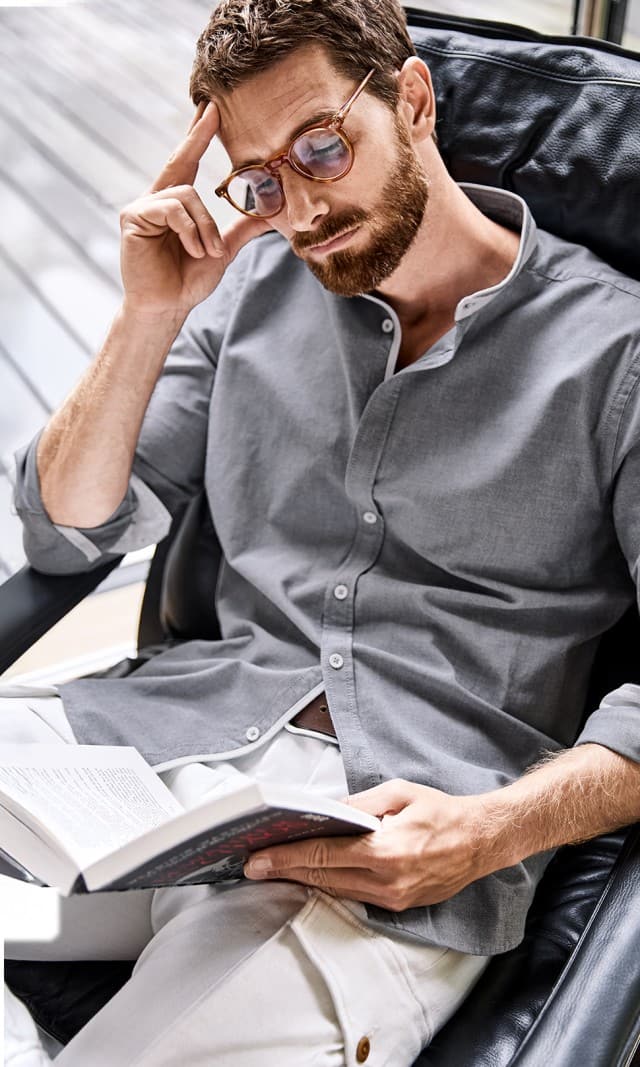 Der Mann trägt ein graues Stehkragenhemd mit hochgekrempelten Ärmeln sowie eine weiße Hose.