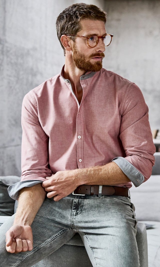 Der Mann trägt ein rosafarbenes Stehkragenhemd mit hochgekrempelten Ärmeln sowie eine hellgraue Jeans.