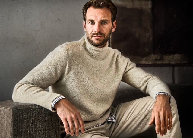 Ein Mann trägt einen dunkelblauen Strickpullover mit hohem Kragen und lehnt entspannt an einer grauen Wand.