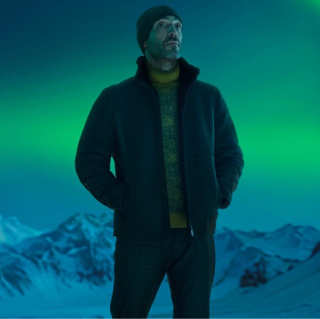 Auf dem Bild steht ein Mann in winterlicher Kleidung vor einer verschneiten Bergkulisse und beobachtet das grün leuchtende Nordlicht am Nachthimmel.