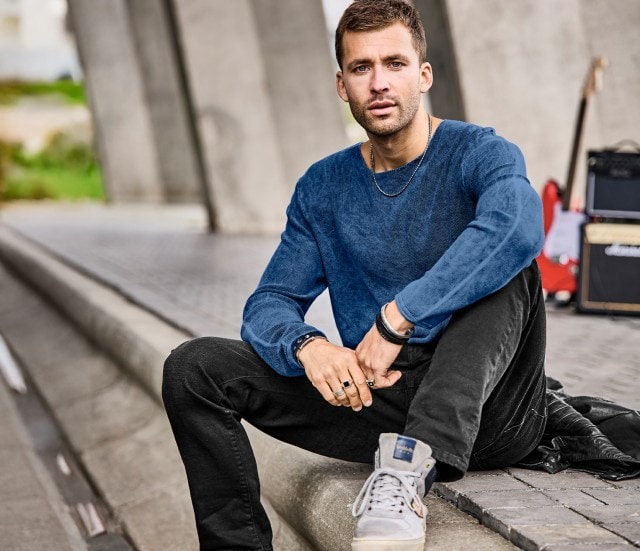 Mann bekleidet mit blauem Pullover und schwarzer Jeans sitzt am Bordstein und blickt in die Kamera