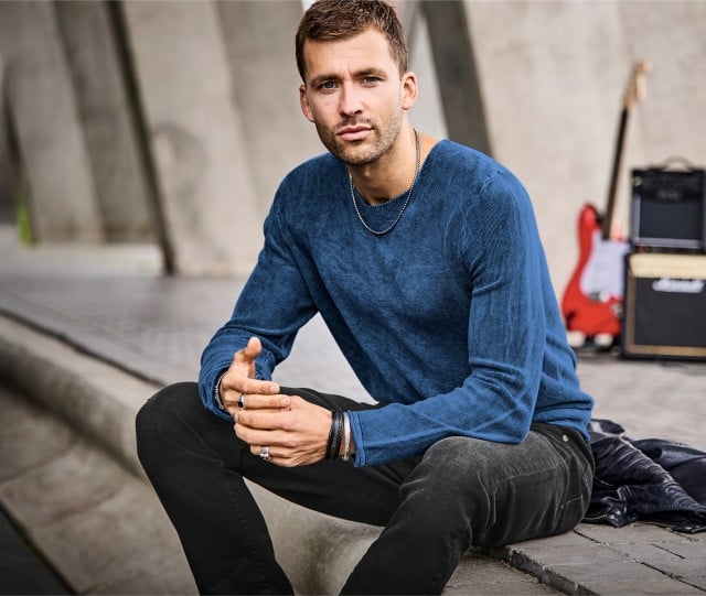 Der Mann trägt einen blauen Strickpullover und eine dunkle, figurbetont geschnittene Hose.
