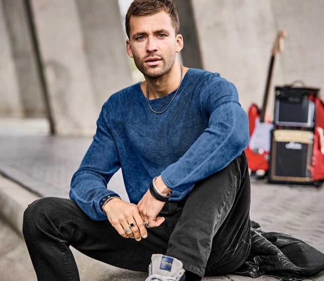 Mann sitzt im Freien und trägt einen blauen Strickpullover mit strukturierter Oberfläche und eine dunkle Hose; rechts daneben ist derselbe blaue Pullover als Produktaufnahme zu sehen.