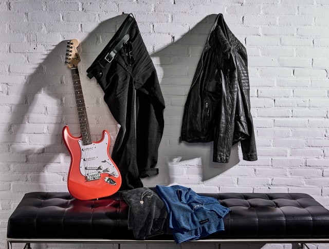 Eine rote E‑Gitarre lehnt an einer weißen Backsteinwand, daneben hängen eine schwarze Jeans mit Gürtel und eine schwarze Lederjacke; auf der schwarzen Sitzbank davor liegen zwei gefaltete Langarmshirts.