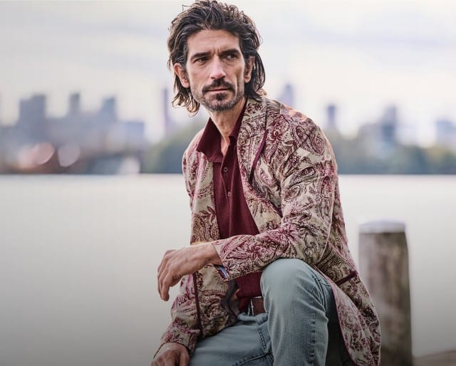 Ein Mann sitzt am Wasser und trägt eine gemusterte beige‑rote Jacke über einem bordeauxfarbenen Shirt sowie eine hellgraue Jeans.