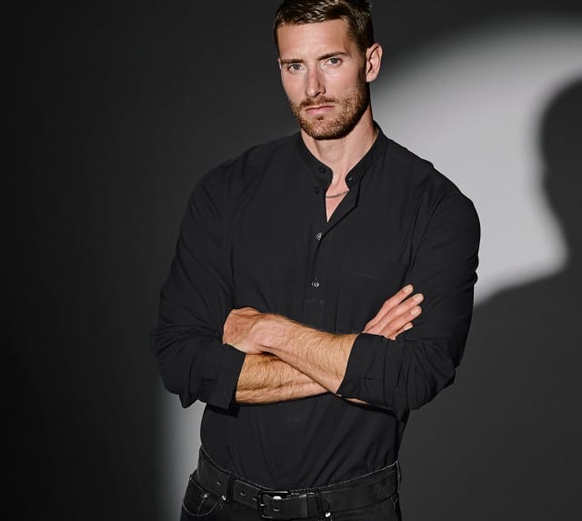 Ein Mann trägt ein schwarzes Stehkragenhemd von Mey & Edlich in Regular Fit, das in einem stilvoll beleuchteten Setting elegant präsentiert wird.