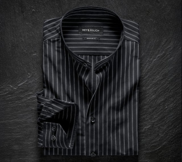Das Bild zeigt ein schwarzes Stehkragenhemd von Mey & Edlich in Regular Fit mit feinen weißen Nadelstreifen, das elegant auf dunklem Untergrund präsentiert wird.