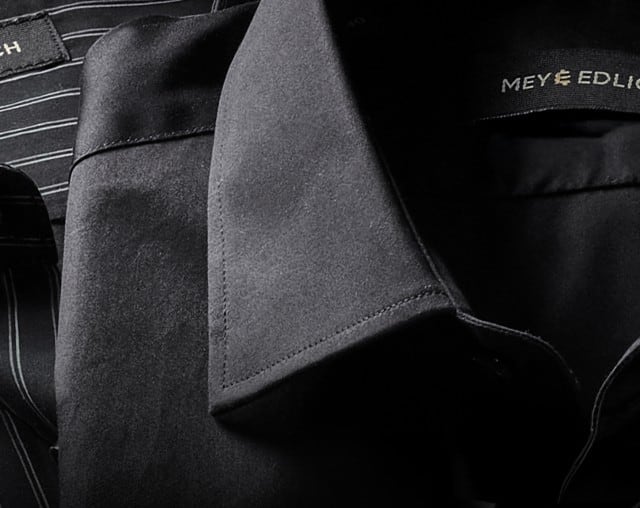 Das Bild zeigt zwei elegante Hemden von Mey & Edlich – eines mit feinen weißen Nadelstreifen und Stehkragen, das andere in schlichtem Schwarz mit klassischem Kragen – stilvoll nebeneinander arrangiert.