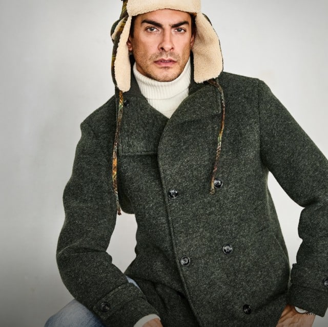 Ein Mann trägt einen dunkelgrünen Wollmantel mit doppelter Knopfreihe, einen cremefarbenen Rollkragenpullover und eine warme Wintermütze mit Ohrenklappen für einen eleganten Winterlook.