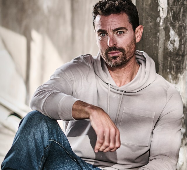 Mann mit einem grauen Hoodie lehnt an einer Wand und schaut in die Kamera
