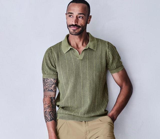 Mann lehnt entspannt an einer hellen Wand und trägt ein grünes Strickpolo