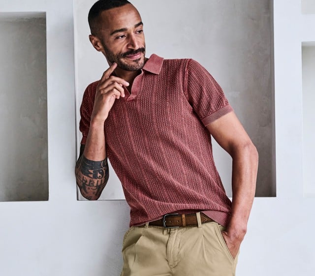 Mann steht schmunzelnd an einer Wand und trägt ein bordeauxfarbenes Polo Shirt und eine beige Hose