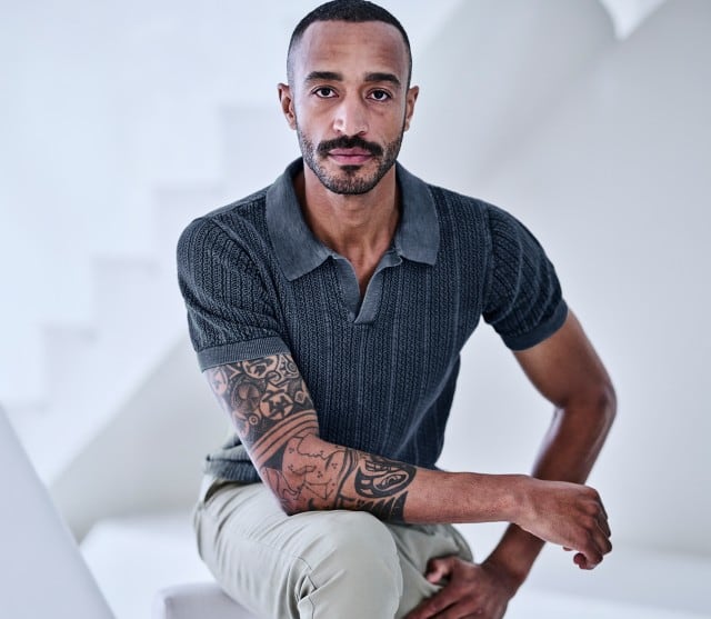 Mann sitzt auf einem Hocker und trägt ein marinefarbenes Strickpolo und eine helle Hose