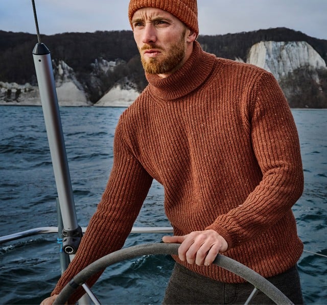 Ein Mann in einem braunen Strickpullover und passender Mütze steuert ein Segelboot auf dem Meer, während im Hintergrund eine Küstenlandschaft mit weißen Kreidefelsen zu sehen ist.