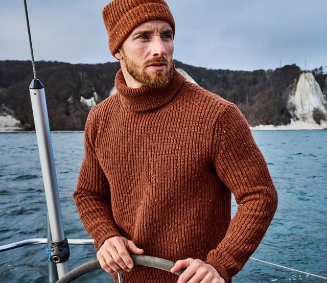 Ein Mann in rostrotem Strickpullover und Mütze steht auf einem Segelboot vor Kreidefelsen, daneben ist eine Nahaufnahme des Wollmaterials zu sehen.