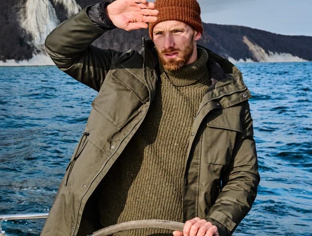 Ein Mann auf einem Segelboot trägt eine olivgrüne Jacke, einen groben Strickpullover und eine braune Mütze, während er sich vor der Sonne schützt, im Hintergrund sind Kreidefelsen zu sehen.