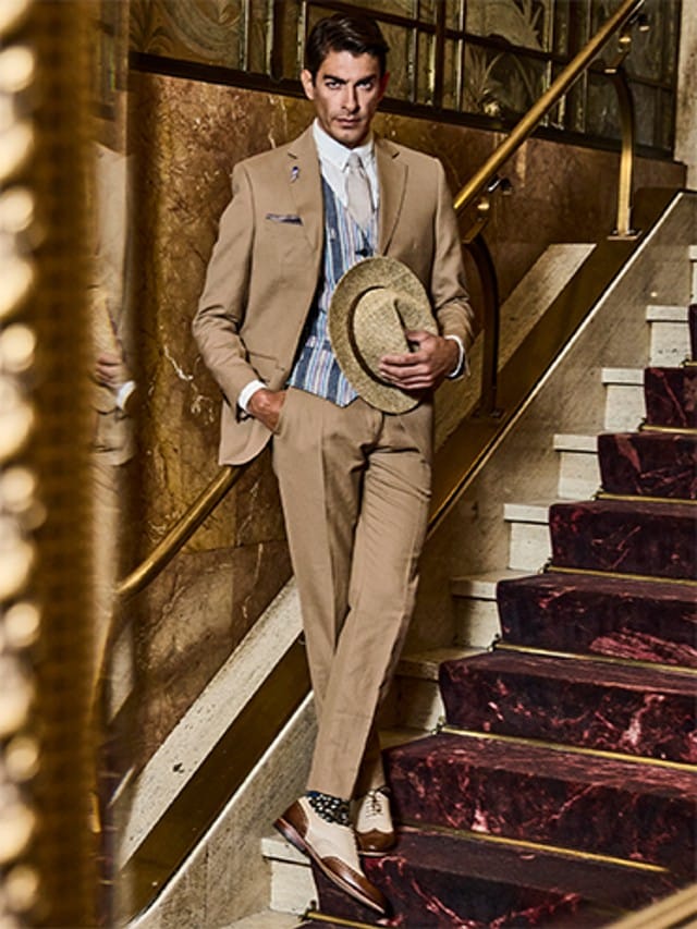Auf dem Bild ist ein elegant gekleideter Mann in einem beigen Anzug zu sehen, der auf einer Treppe steht und einen Strohhut in der Hand hält.