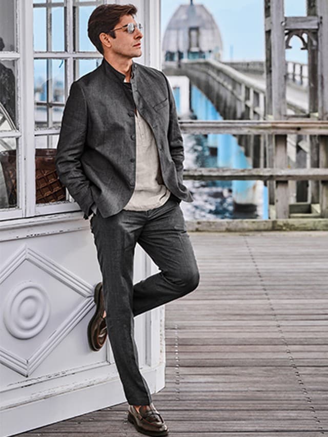 Auf dem Bild ist ein Mann zu sehen, der lässig in einem stilvollen grauen Anzug mit beigem Pullover und Sonnenbrille an einer Holzhütte auf einem Pier steht.


